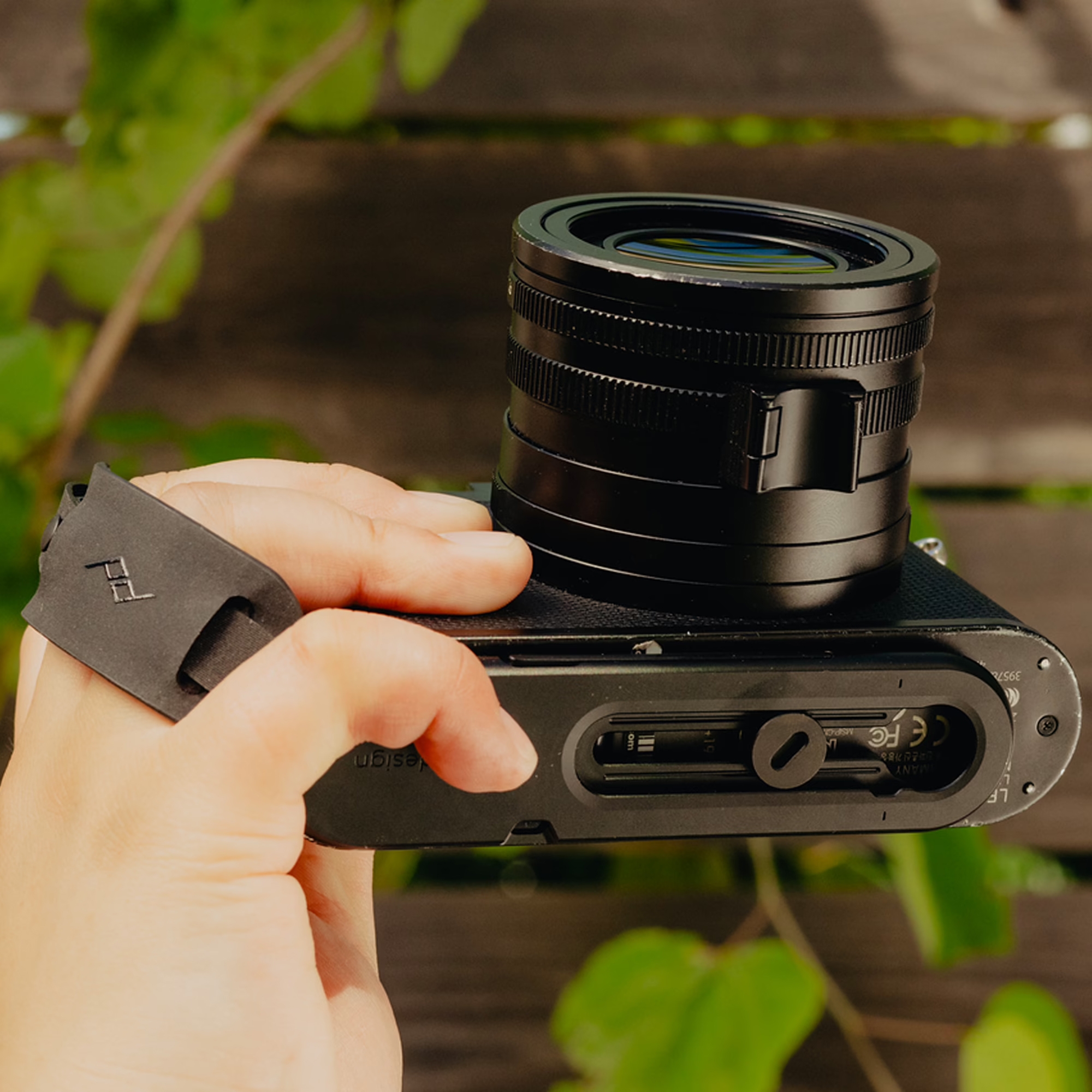 Eine Hand hält eine schwarze Leica Kamera von unten, um die montierte Peak Design Micro Clutch Handschlaufe samt Bodenplatte und integriertem Werkzeug in der Praxisanwendung vor einem unscharfen, grünen Hintergrund zu zeigen.